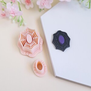May include: Three pink plastic cookie cutters shaped like spiderwebs. The largest cutter has the word "WEB" written on it. The smallest cutter has the word "WEB" written on it. The middle cutter has the word "CCC" written on it. A black plastic spiderweb cookie cutter with a purple centre is on a white surface.