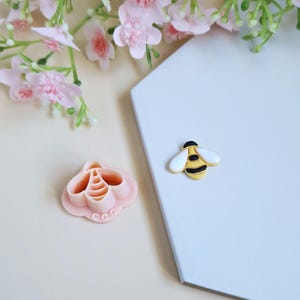Peut inclure: Un emporte-pièce rose en forme d'abeille et une petite figurine d'abeille jaune, noire et blanche sont présentés. L'emporte-pièce est sur une surface beige clair, tandis que la figurine d'abeille repose sur une surface blanche. Des fleurs roses sont en arrière-plan.