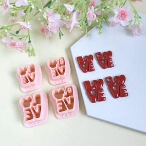 Könnte beinhalten: Rosa Ausstecher und rote "LOVE" Verzierungen sind auf einer weißen Oberfläche angeordnet, mit rosa Blumen im Hintergrund. Die Ausstecher haben die Form des Wortes "LOVE".