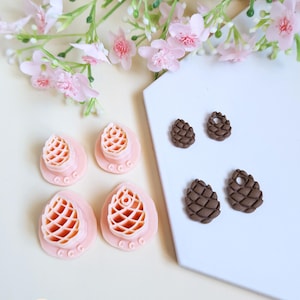 Peut inclure: Des découpoirs à biscuits roses et des boucles d'oreilles en forme de pommes de pin marron sont présentés sur une surface blanche, avec des fleurs roses en arrière-plan. Les découpoirs ont un motif de pomme de pin.