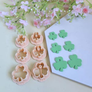 Könnte beinhalten: Eine Sammlung von Kleeblatt-Ausstechern und grünen Kleeblatt-Ausschnitten. Die Ausstecher sind hellrosa und die Ausschnitte sind grün. Rosa Blüten im Hintergrund deuten auf ein St. Patrick's Day-Thema hin.