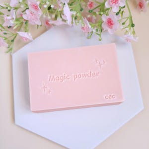 Puede incluir: Una barra de jabón rosa con el texto "Magic powder" y "CCC" en relieve. El jabón está sobre una superficie hexagonal blanca.