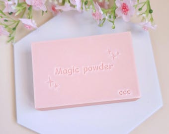 Puede incluir: Una barra de jabón rosa con el texto "Magic powder" y "CCC" en relieve. El jabón está sobre una superficie hexagonal blanca.