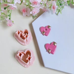 May include: Pink heart-shaped cookie cutters and heart-shaped earrings with gold star accents. The cookie cutters have a scalloped edge. The earrings are on a white surface with pink flowers in the background.