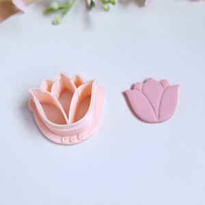 Op de afbeelding: Een roze plastic koekjesvormpje in de vorm van een bloem en een roze fondantbloem. De koekjesvormpje heeft een gedetailleerd ontwerp en de fondantbloem is licht verhoogd.