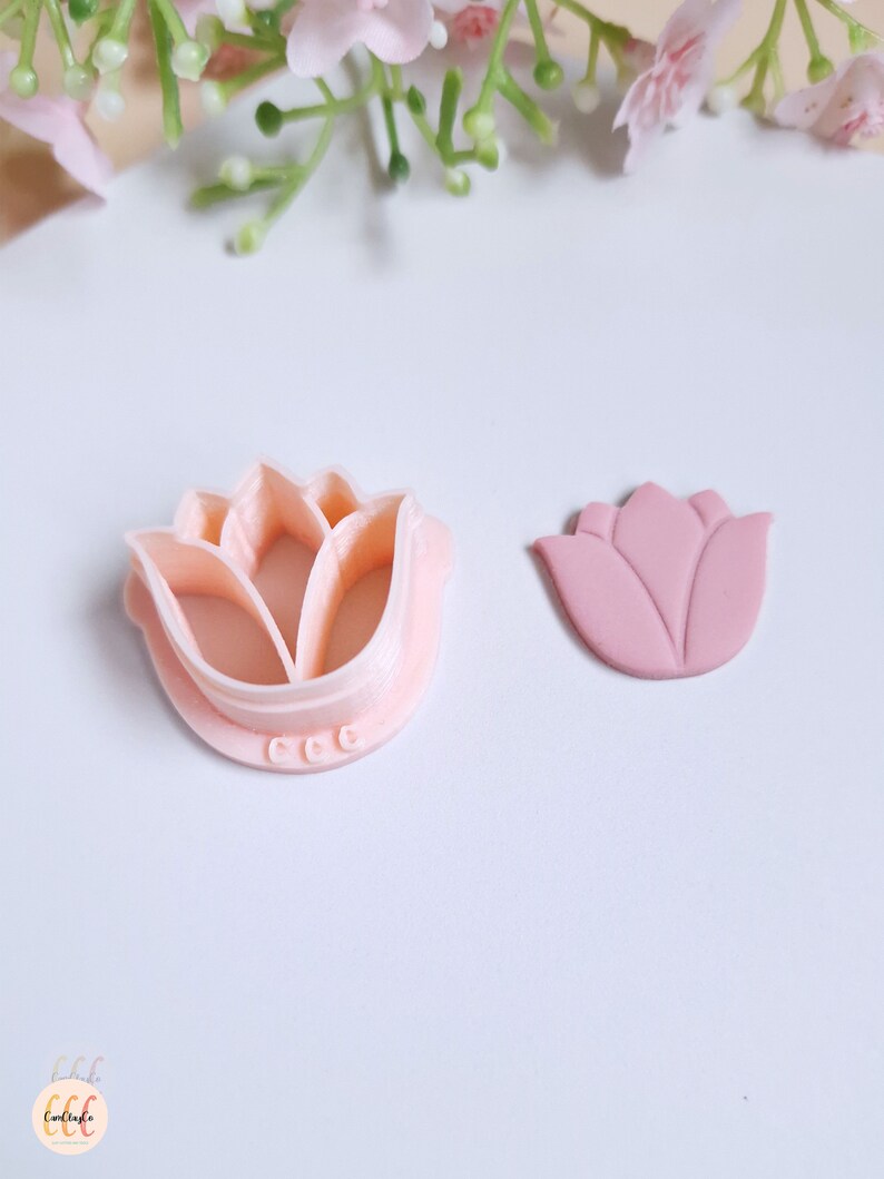 Op de afbeelding: Een roze plastic koekjesvormpje in de vorm van een bloem en een roze fondantbloem. De koekjesvormpje heeft een gedetailleerd ontwerp en de fondantbloem is licht verhoogd.