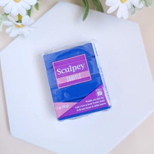 Puede incluir: Un bloque azul de arcilla polimérica de la marca Sculpey Souffle, envasado en una envoltura de plástico transparente. El paquete tiene una etiqueta morada con el nombre de la marca, el nombre del producto y el peso: 1,7 oz (48 g). La etiqueta también incluye el texto "Premium Oven-Bake Clay" en inglés, francés y español.