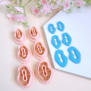 Puede incluir: Un juego de seis cortadores de galletas de plástico rosa en forma de corazón con un agujero en el medio. Los cortadores están etiquetados con los números 2002, 2003, 2004, 2005, 8002 y 88. Los cortadores están dispuestos en círculo sobre una superficie blanca. También hay cinco cortadores de galletas de plástico azul en forma de línea ondulada con un agujero en el medio sobre una superficie blanca.
