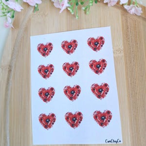 Può includere: Un foglio di adesivi con disegni a forma di cuore rosso con illustrazioni di fiori di papavero. Gli adesivi sono disposti su uno sfondo bianco. Il marchio "CamClayCo" è stampato in basso a destra. L'immagine è su uno sfondo di legno.