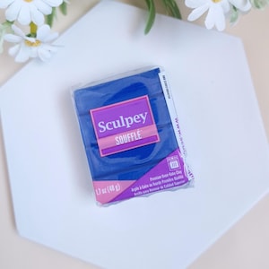 Peut inclure: Un paquet de pâte polymère Sculpey Soufflé bleu, avec le nom de la marque et du produit en rose et violet. Le paquet pèse 48 grammes.