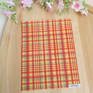 May include: A rectangular sheet with a red and yellow plaid pattern, with thin green lines. The sheet has the text "CamClayCo" in the bottom right corner. The sheet is on a wooden surface, with pink flowers in the background.