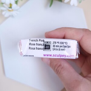 May include: A rectangular package of French Pink polymer clay. The label includes the text "French Pink", "275 &deg;F (130 &deg;C)", and "www.sculpey.com". The clay is held in a hand, with a white surface and flowers in the background.
