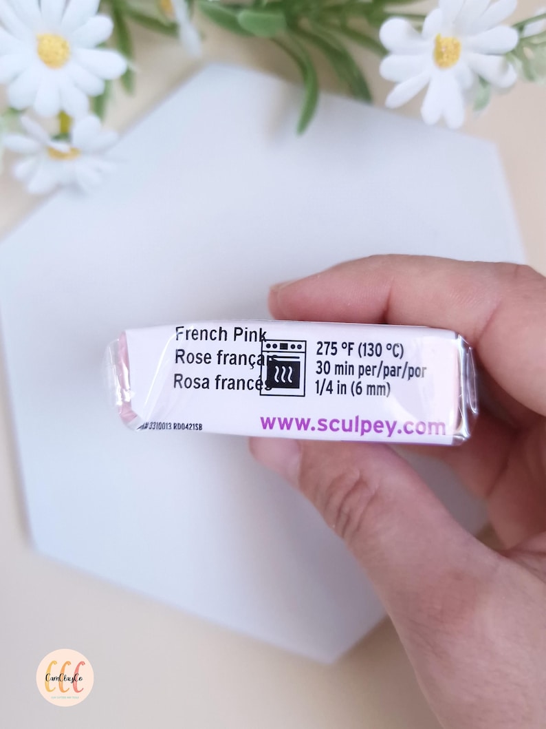 May include: A rectangular package of French Pink polymer clay. The label includes the text "French Pink", "275 &deg;F (130 &deg;C)", and "www.sculpey.com". The clay is held in a hand, with a white surface and flowers in the background.
