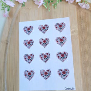 Könnte beinhalten: Ein Bogen Aufkleber mit herzförmigen Motiven, gefüllt mit roten Rosen und kleinen rosa Blüten. Die Aufkleber haben einen weißen Hintergrund und sind in Reihen und Spalten angeordnet. Der Markenname "CamClayCo" ist unten rechts aufgedruckt.