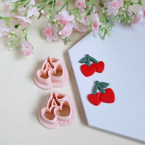 Peut inclure: Des découpoirs à biscuits roses en forme de cerise et deux boucles d'oreilles cerises rouges avec des tiges et des feuilles vertes sont présentés sur une surface blanche. Le fond présente des fleurs roses. Les découpoirs ont des découpes en forme de cœur.