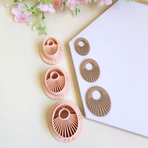 May include: Three light pink plastic clay cutters in graduated sizes, each featuring a circular center and radiating fan-like lines. Beside them are three corresponding brown clay shapes with the same sunburst pattern. The cutters are marked 20, 25, and 30.
