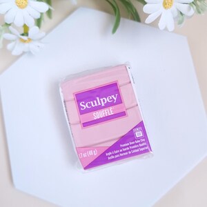 May include: A package of pink Sculpey Souffl&eacute; polymer clay, 1.7 oz (48 g), with the product name in a purple rectangle. The package also includes text in French and Spanish. The clay is on a white hexagonal surface, with white flowers in the background.
