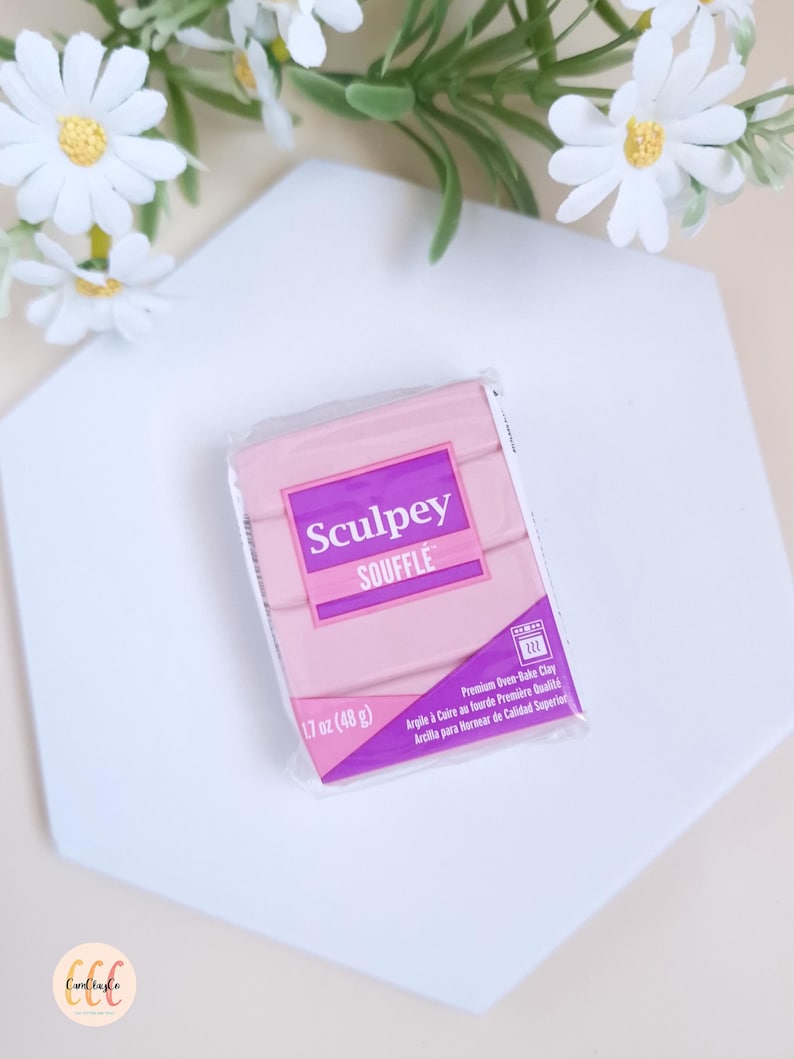 May include: A package of pink Sculpey Souffl&eacute; polymer clay, 1.7 oz (48 g), with the product name in a purple rectangle. The package also includes text in French and Spanish. The clay is on a white hexagonal surface, with white flowers in the background.