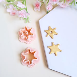 Può includere: Due formine rosa a forma di fiore e due decorazioni dorate a forma di stella sono esposte. Le formine hanno un centro a forma di stella. Fiori rosa sullo sfondo.