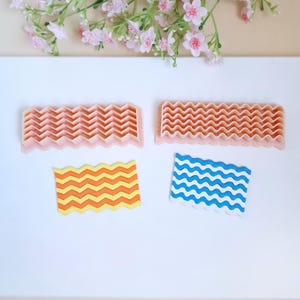 Puede incluir: Dos herramientas de decoración de pasteles de plástico rosa con patrones ondulados. Las herramientas se muestran junto a dos muestras de decoración de pasteles, una con líneas onduladas amarillas y naranjas y la otra con líneas onduladas azules y blancas.