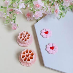 May include: Pink flower-shaped cookie cutters and flower-shaped embellishments with red heart centres. The cutters have a detailed design, and the embellishments are on a white surface, with pink flowers in the background.
