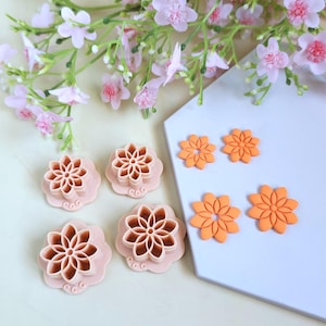 Peut inclure: Ensemble de découpoirs en argile en forme de fleurs dans des tons pêche et orange, avec des fleurs en argile assorties. Les découpoirs présentent des motifs de pétales complexes, et les fleurs en argile sont de différentes tailles. Fleurs roses et verdure en arrière-plan.