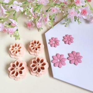 Könnte beinhalten: Eine Sammlung rosa blütenförmiger Ton-Ausstecher und blütenförmiger Tonverzierungen. Die Ausstecher sind in verschiedenen Größen erhältlich und weisen detaillierte Blütenblattdesigns auf. Rosa Blumen und Grünzeug im Hintergrund.