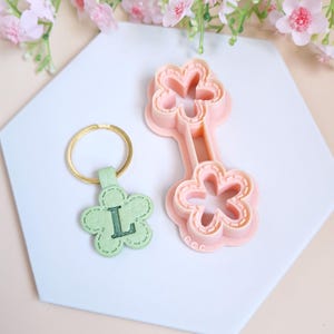 May include: A pink plastic flower-shaped cookie cutter with a smaller flower-shaped cookie cutter attached to the top. A green leather keychain with a flower shape and the letter "L" is also shown.