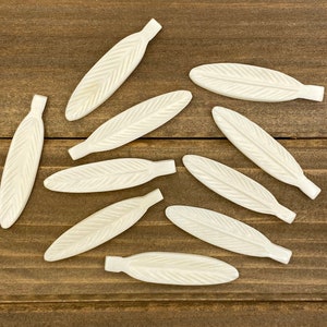 May include: A collection of ten white, carved, feather-shaped ornaments on a brown wooden surface.