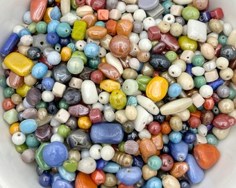 May include: A white bowl filled with a variety of colorful ceramic beads. The beads are in various shapes and sizes, including round, oval, square, and rectangular. The colors include blue, green, red, orange, yellow, brown, and white.