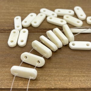 May include: Ivory-colored rectangular beads with two holes on each end, strung together with clear thread. The beads are scattered on a wooden surface, some in rows and others loose. The beads have a smooth, slightly glossy finish.