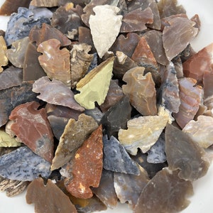 May include: A collection of arrowheads made from various types of stone, including agate, jasper, and flint. The arrowheads are in a variety of colors, including brown, red, green, and gray. They are all in a bowl, and some are stacked on top of each other.