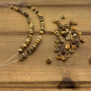 May include: Strung beads in shades of brown, tan, and cream, scattered on a wooden surface. Some beads are strung into a necklace, while others are loose. The beads have a natural, earthy appearance, suggesting a handmade jewelry component.