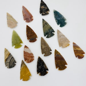May include: A collection of fifteen stone arrowheads in various colors and patterns. The arrowheads display a range of hues, including beige, red, green, brown, and black. Each arrowhead has a distinct shape and texture, showcasing natural stone variations.