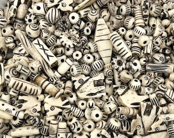 May include: A white bowl filled with a variety of bone beads. The beads are all different shapes and sizes, and some have intricate designs carved into them.
