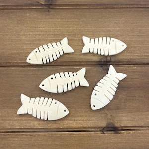 May include: Five small, off-white fish-shaped wooden cutouts with etched details, arranged on a brown wooden surface. Each fish has a tail fin, eye, and rib-like markings.
