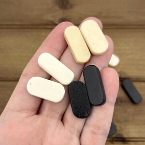 May include: A hand holding a collection of small, rectangular game pieces. The pieces are in various colors, including cream, black, and off-white. The wooden pieces have rounded edges and a smooth finish. The background is a wooden surface.