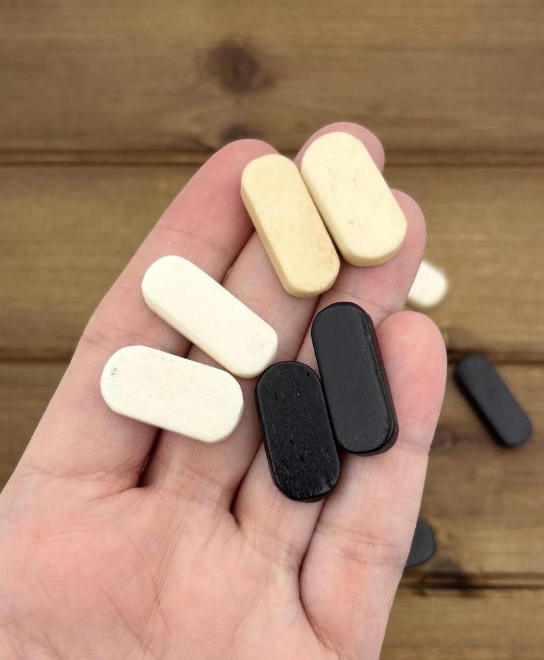 May include: A hand holding a collection of small, rectangular game pieces. The pieces are in various colors, including cream, black, and off-white. The wooden pieces have rounded edges and a smooth finish. The background is a wooden surface.
