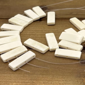 May include: Ivory-colored rectangular beads with three holes are arranged on a wooden surface. The beads are strung on a clear, thin cord. The beads have a slightly aged appearance, suggesting a vintage or natural material.