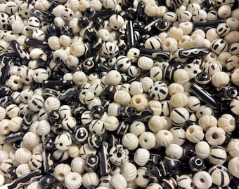 May include: A collection of ivory and black beads of various shapes and sizes. The beads include round, cylindrical, and patterned designs. The ivory beads have a natural, off-white color, while the black beads are a deep, solid black. The beads are scattered, creating a textured surface.