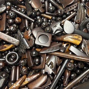 May include: A close-up shot of a collection of various dark brown and silver-toned beads and charms. The assortment includes star shapes, square shapes, and cylindrical beads. Some pieces have a wood-like appearance, while others are metallic.