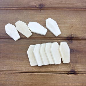 May include: Ten wooden coffin shaped ornaments in a light beige color. The ornaments are arranged in two rows on a wooden surface.