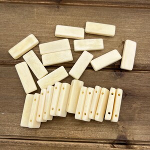 May include: Ivory-colored rectangular beads with rounded edges, some with multiple holes for stringing. The beads are scattered on a wooden surface, showcasing their smooth texture and natural hue. Ideal for jewelry making or crafting.