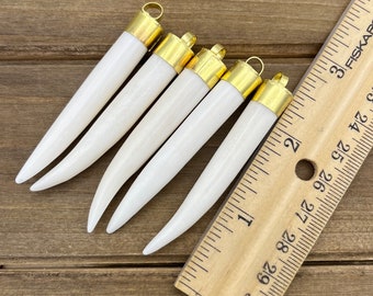 May include: Five white, tooth-shaped pendants with gold-colored metal caps. The pendants are arranged in a fan shape and are measured against a wooden ruler.