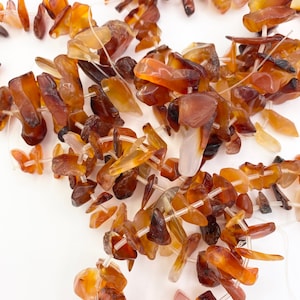 Op de afbeelding: Een streng carneol edelsteen kralen, elke kraal heeft een unieke, onregelmatige vorm met een roodbruine kleur. De kralen zijn geregen op een doorzichtig elastisch koord.