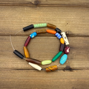 Könnte beinhalten: Eine Halskette mit einer Vielzahl von bunten, zylindrischen Perlen. Die Perlen bestehen aus verschiedenen Materialien und Farben, darunter Schwarz, Grün, Orange, Blau und Holztöne. Die Halskette wird auf einer Holzoberfläche präsentiert.