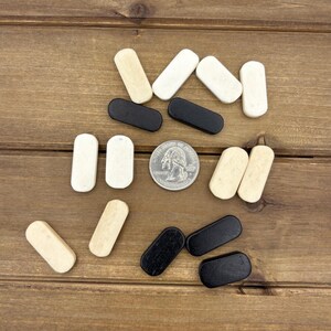 May include: A collection of small, rectangular wooden game pieces in black, white, and cream colors. A silver coin is also present. The pieces are arranged on a brown wooden surface, suggesting a game or craft supply.