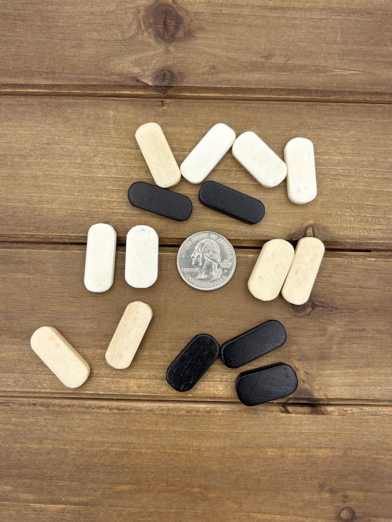 May include: A collection of small, rectangular wooden game pieces in black, white, and cream colors. A silver coin is also present. The pieces are arranged on a brown wooden surface, suggesting a game or craft supply.