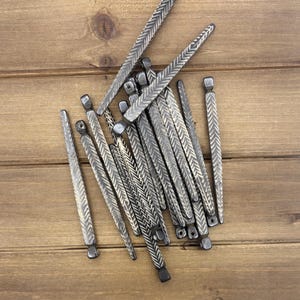 May include: A collection of silver-toned metal hair sticks with a textured, chevron-like pattern. The hair accessories are scattered on a wooden surface, showcasing their decorative design and potential for hairstyling.