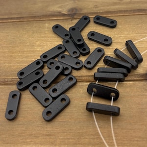 May include: A collection of small, black, rectangular wooden beads with two holes each. The beads are scattered on a light brown wooden surface. Some beads are strung together with white string.